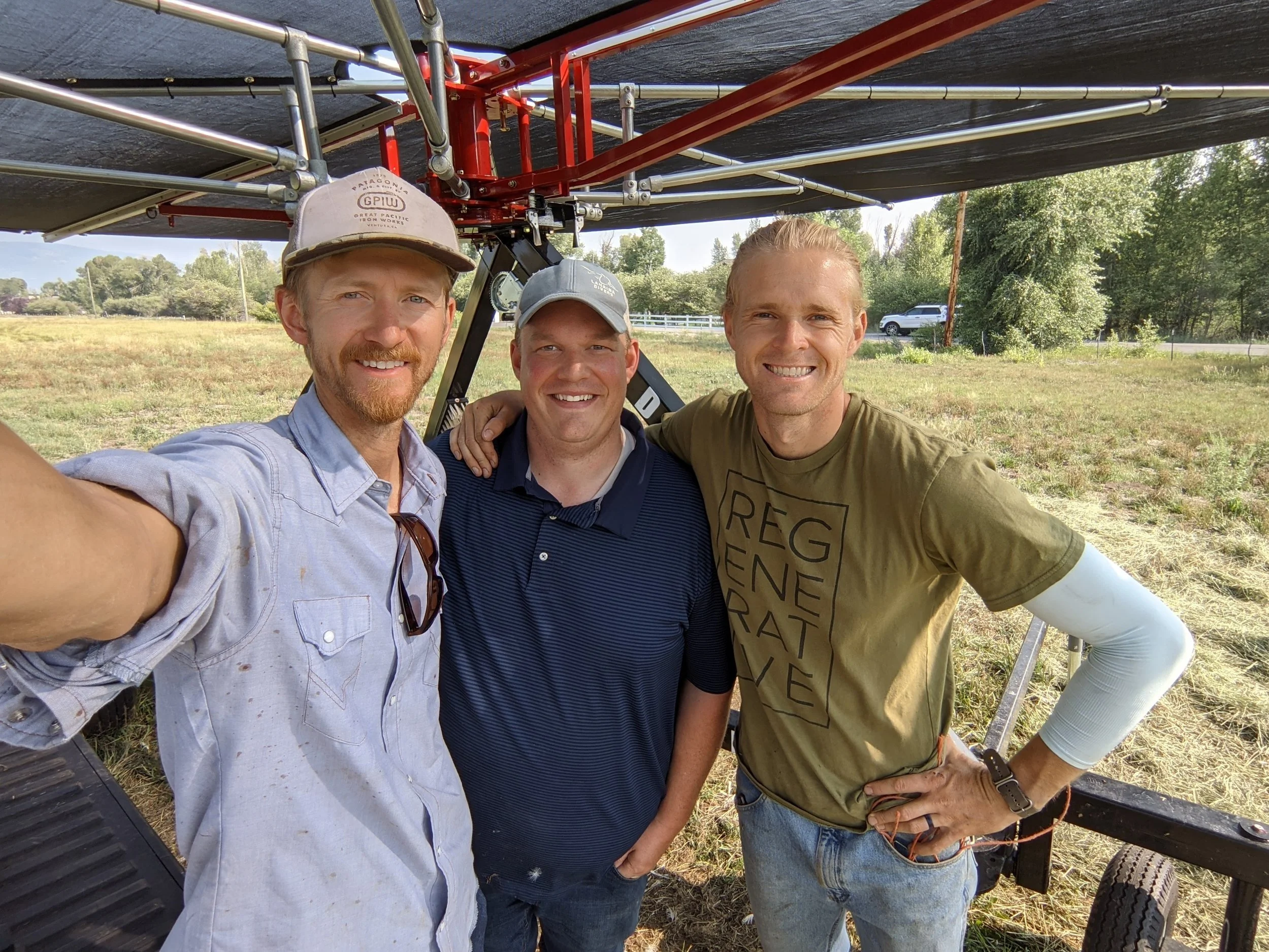 Three men smiling for a selfie in a grassy field, standing under a large shaded structure. They are dressed casually, with one wearing a light blue shirt, another in a navy polo, and the third in a green shirt with "REGEN" printed on it. The background features trees and a vehicle on a dirt road.