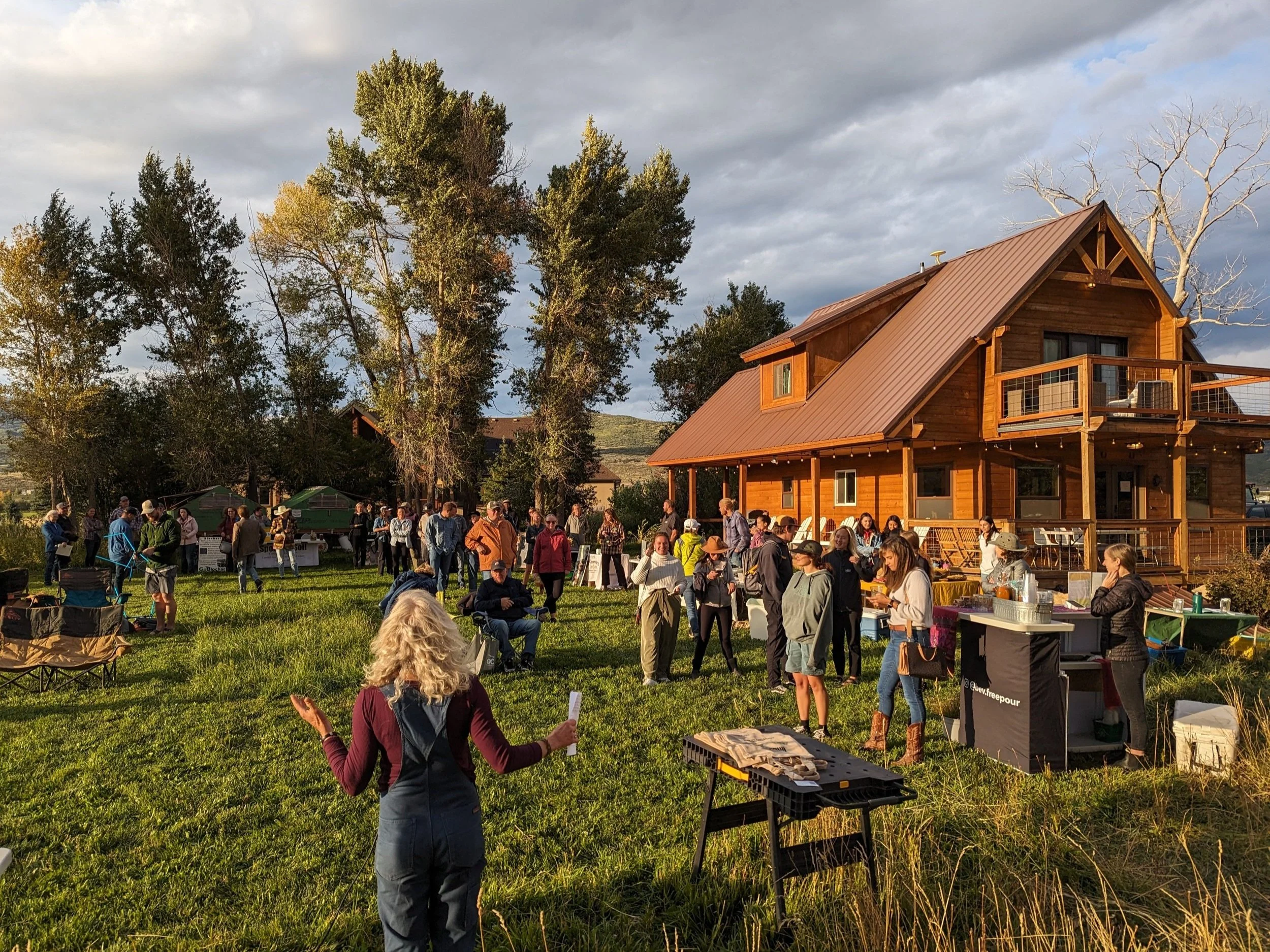 Outdoor gathering at a rustic wooden cabin, featuring a diverse group of people socializing, enjoying food and drinks, with tall trees and a scenic landscape in the background. The event showcases a lively atmosphere, perfect for community bonding and celebration.