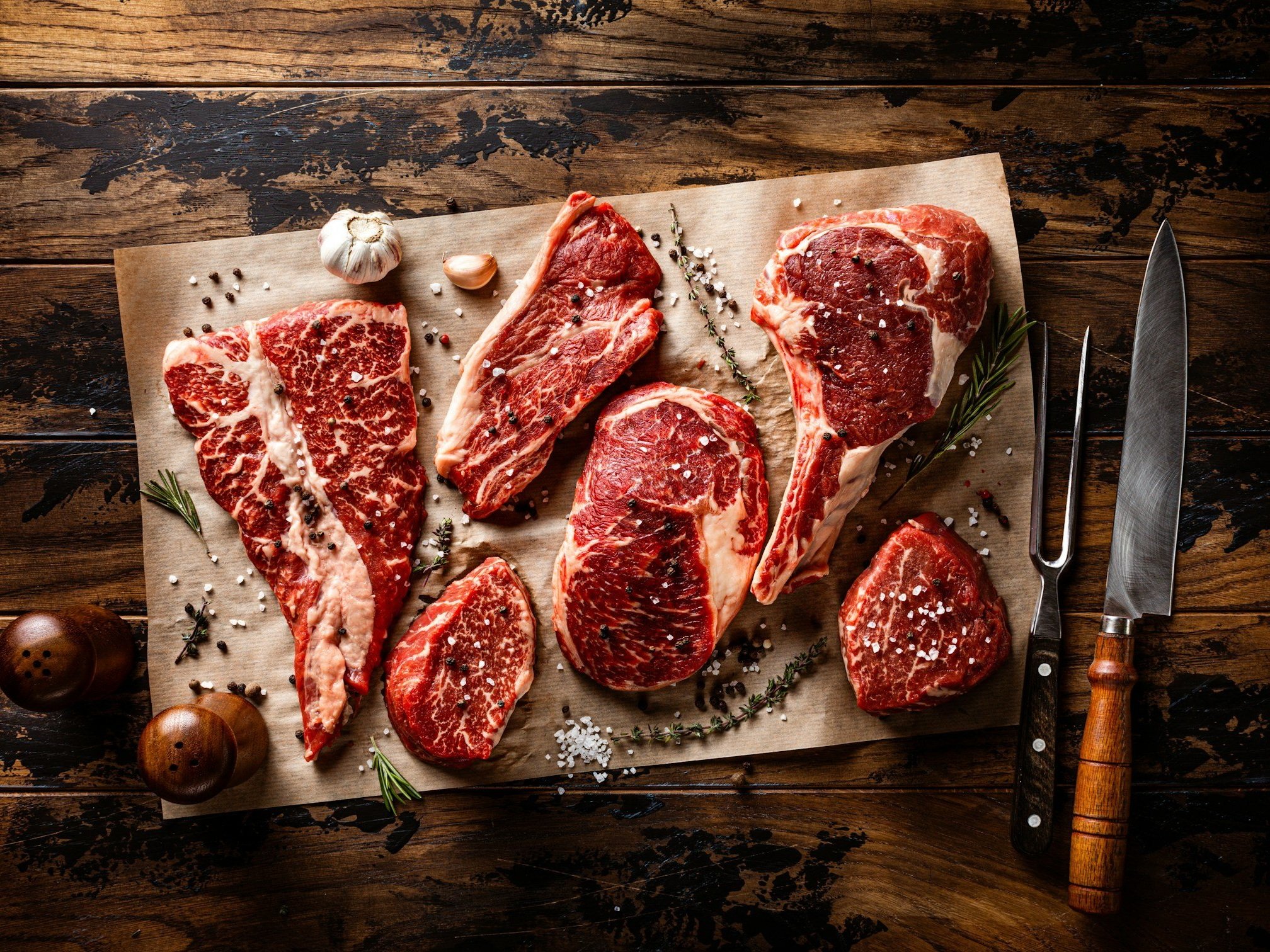 unsplash-image-JHWfH8V2INk (2) Assorted cuts of marbled beef arranged on butcher paper, garnished with herbs and spices, alongside a knife and fork on a rustic wooden surface. Ideal for cooking enthusiasts and meat lovers.