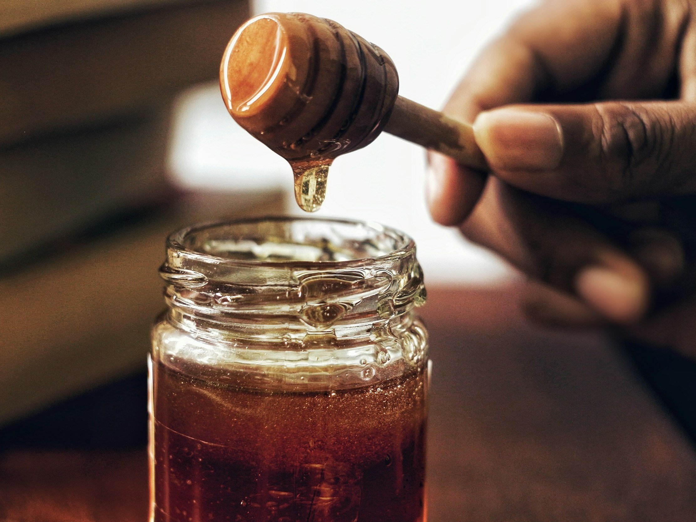 unsplash-image-yQzrDgU-KAI Close-up of a hand holding a honey dipper dripping honey into a glass jar, showcasing the rich texture and color of the honey against a blurred background of books. Ideal for topics related to natural sweeteners, cooking, or health benefits of honey.